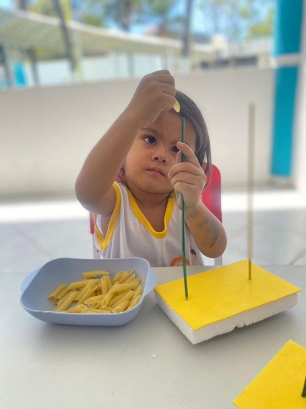 Criança encaixando macarrão em palitos numa atividade de coordenação motora